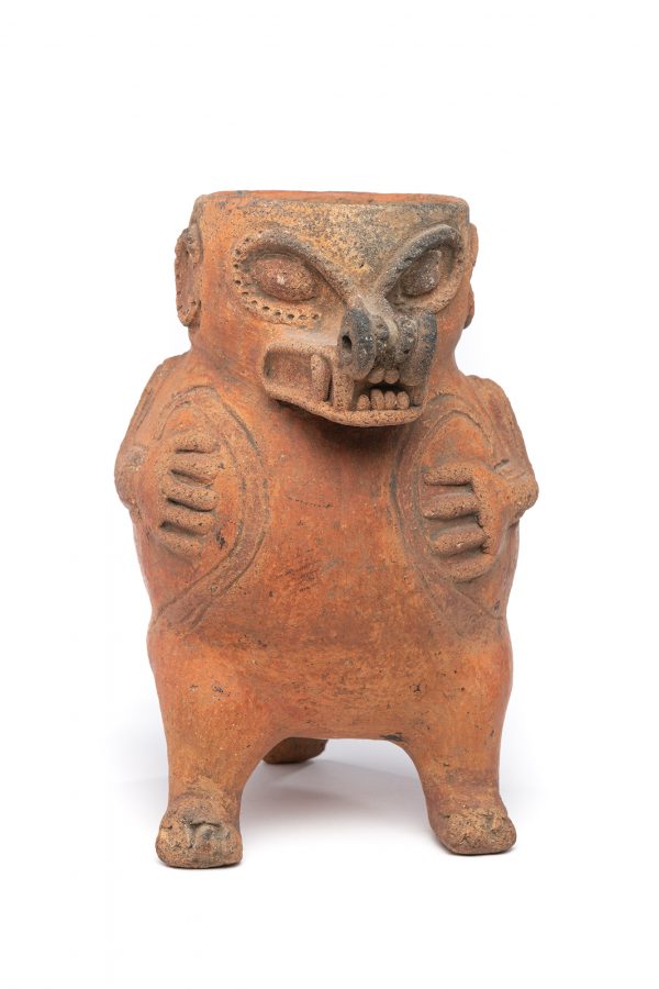 A jar with the two feet and tail as the tripod foot of the jar. The middle of the jar has projecting arms and hands, the neck of the jar has a projecting nose of the animal. The nose and around the eyes has a decorative rim with holes, over a mouth with teeth and fangs.