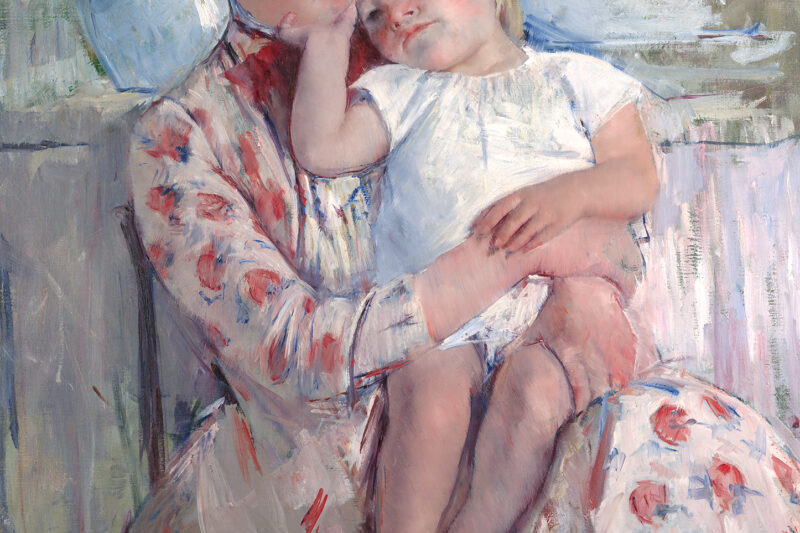 A woman in a flowered dress holds a child. There is a blue vase and wash basin behind her.