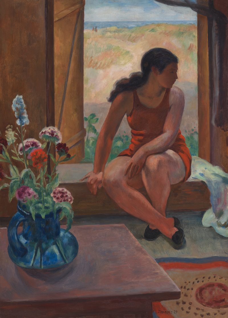 A woman sits before a window facing indoors but looking to her left. There is a blue vase with flowers on the left side.