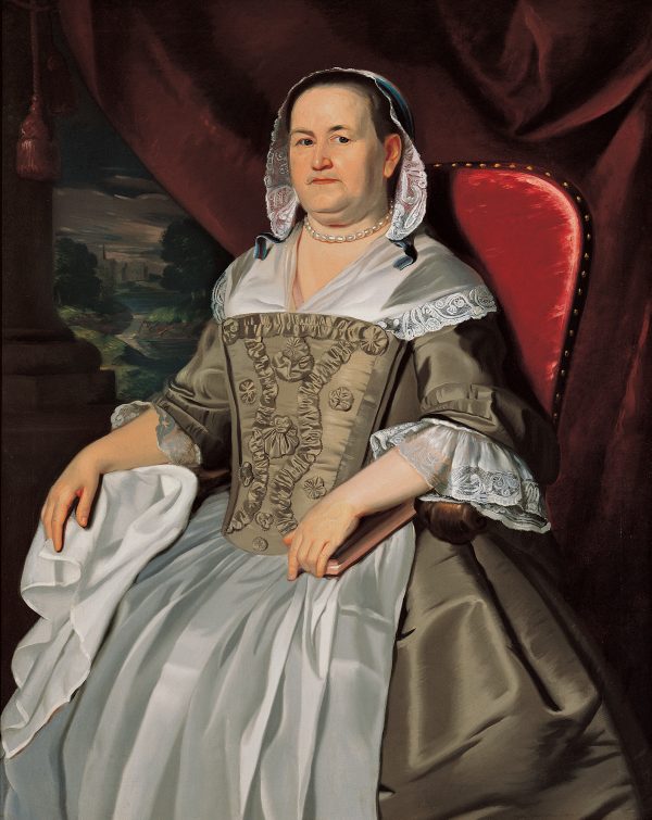 A woman seated on a red chair. She holds a closed book and wears a lace covering over her hair.