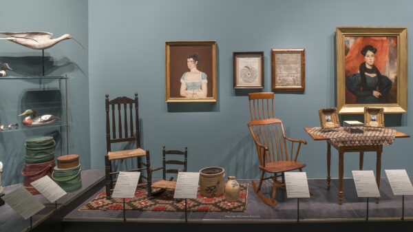 Photo of early American decorative arts includes two chairs, one rocking chair, two paintings of somen, a desk, framed photos, a rug, a milk jub, a crock and a duck decoy