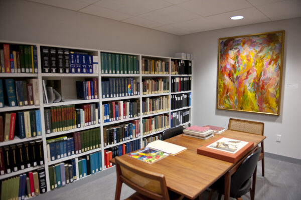 Interior view of the Library with a table, three chairs, books on the table, shelves filled with books in the background and a colorful abstract painting on the wall