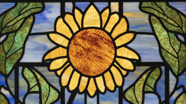 Stained glass window with a yellow sunflower in the center surrounded by green leaves with a blue sky in the background