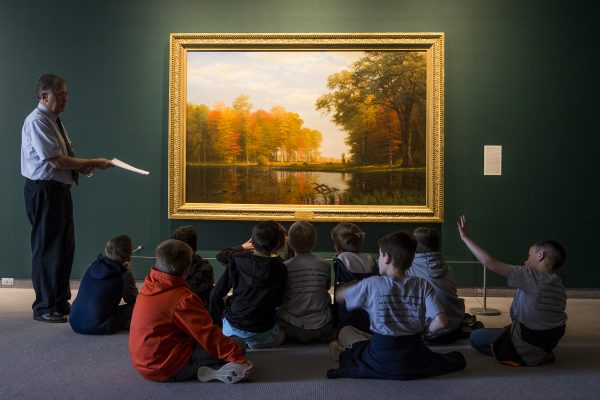 Interior view of galleries as an adult is standing in front of a landscape painting speaking to a group of students on a field trip tour