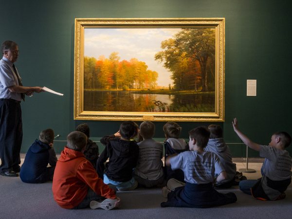 Interior view of galleries as an adult is standing in front of a landscape painting speaking to a group of students on a field trip tour