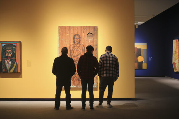 Photo of three men in shadow standing in front of a large painting of a man and a woman