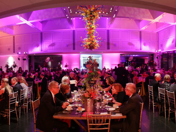 Photo of dozens of people sitting at decorated tables and chairs eating dinner in the Farha Great Hall with the large Chihuly Chandelier in the top center