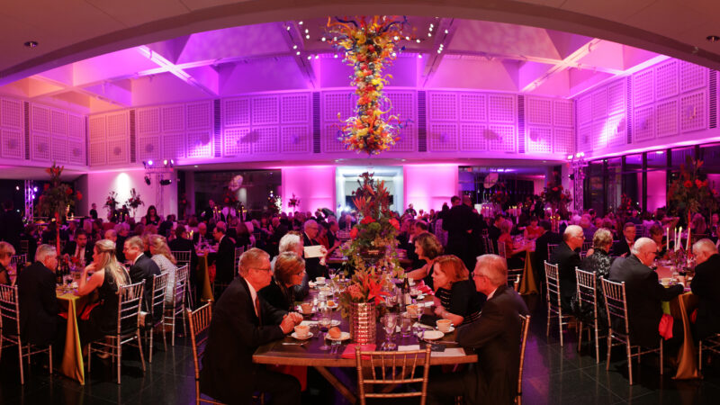 Photo of dozens of people sitting at decorated tables and chairs eating dinner in the Farha Great Hall with the large Chihuly Chandelier in the top center