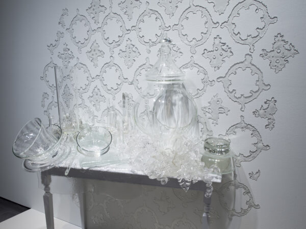 Detail of a clear glass sculpture of vases and other vessels laid on a white table with ornate glass wallpaper on the wall behind the table