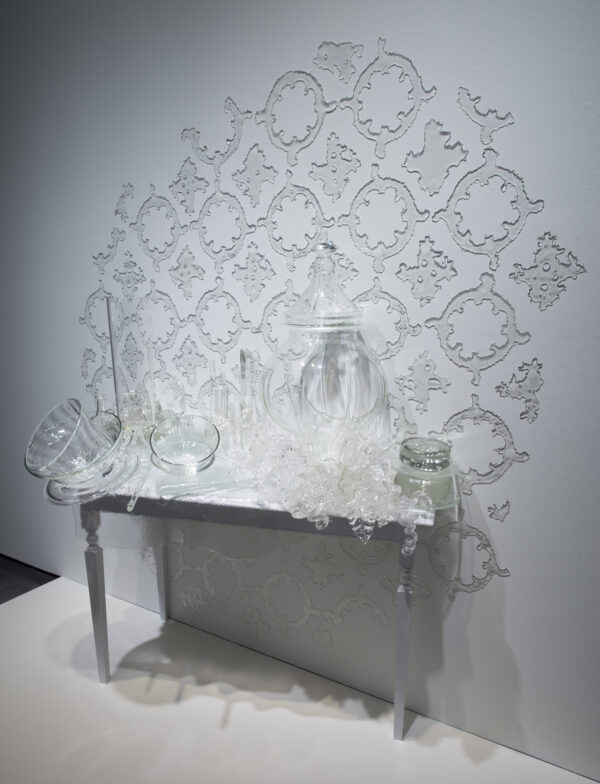 Detail of a clear glass sculpture of vases and other vessels laid on a white table with ornate glass wallpaper on the wall behind the table