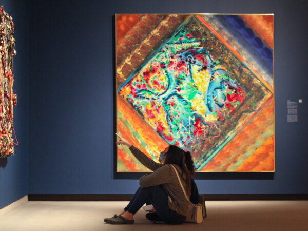 Photo of two people sitting on the floor in front of a colorful abstract painting, with one pointing to another painting on the all in front of them.