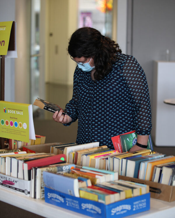 Woman in a face mask, looking at the back of a paperback book, standing in front of a table of books