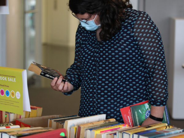 Woman in face mark holding a paperback book in her hand, standing at a table of books.