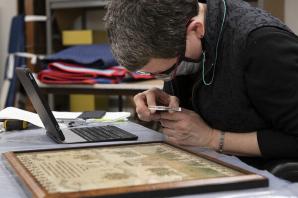 woman wearing a face mask uses a magnifying glass to examine a framed, embroidered sampler.