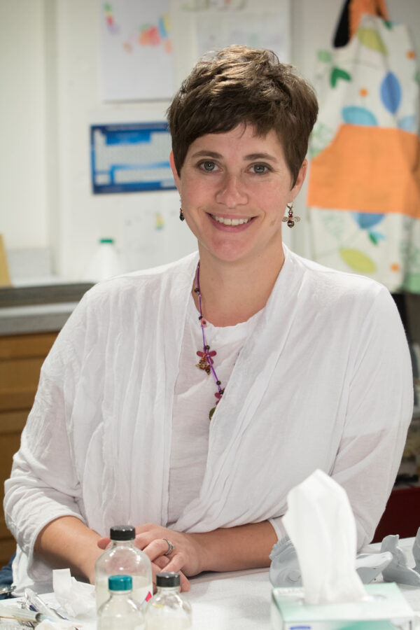 Woman with short hair and a white shirt with red earrings and red necklace looks directly at the camera with clear bottles and a box of facial tissues in front of her and a wall of printouts behind her