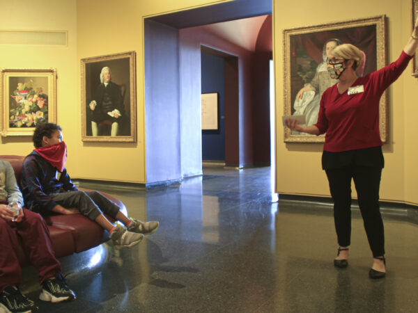 Interior of the galleries with a woman on the right gesturing toward a painting and two young men on the left sitting and watching