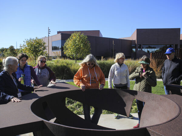 Photograph of a group of people looking at a large-scale circular sculpture of weathered metal in the museum art garden with the museum building in the background