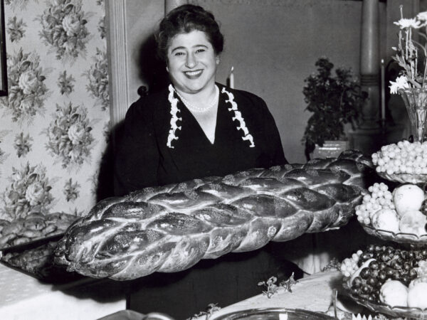 Smiling woman sitting at a table with a large challah bread in front of her and a three-tiered serving bowl with grapes and other fruit and topped with cut flowers