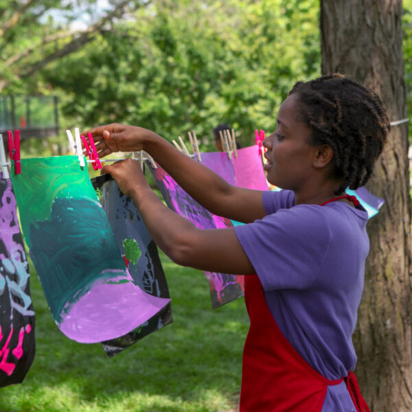 Girl in purple shirt and red apron hanging an anstract artwork on a string tied to a tree in the Art Garden