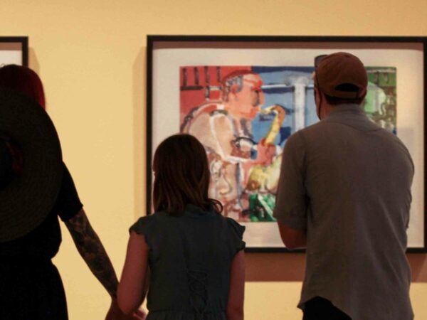 Three people, adult woman holding the hand of a young girl and adult man, in silhouette in front of colorful painting of a saxophone player in a gallery