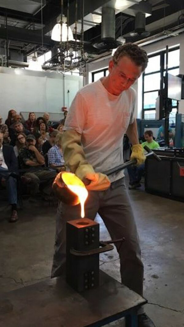 Photograph of a man pouring molten glass into a mold in front of an audience.