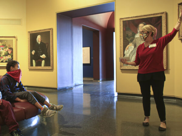 Woman on the right in the galleries gesturing towards a painting while two boys on the left sit on a round sofa and watch