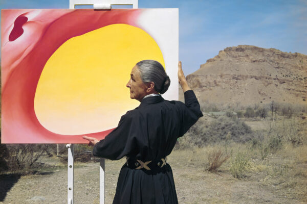 Photograph of artist Georgia O'Keeffe standing with her back to the photographer, holding a large painting of swirls of orange and yellow. She is dressed in a long-sleeved black dress with a leather belt with criss-crosses and her dark hair with gray is in a tight low bun. The landscape behind the painting is a desert setting with a mountain in the background.