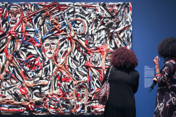 Two women with their backs toward the camera looking at a multi-colored abstract painting against a blue gallery wall