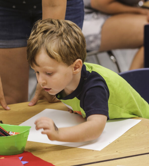 Photograph of a young boy selecting a crayon from a green plastic basket on the table in front of him. He has light brown hair and is dressed in a lime green t-shirt with black sleeve and collar.