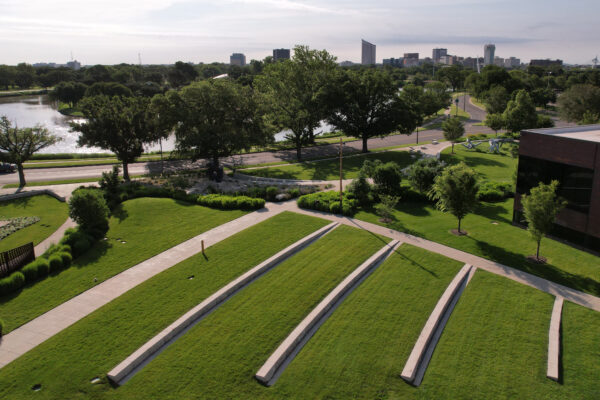 Aerial photograph of the museum's east side in the Art Garden and showing three long slabs of limestone benches on green grass in the Paula and Barry Downing Amphitheater with the City of Wichita skyline and half-dozen buildings in the background