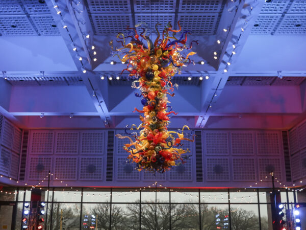 Photo of the colorful, large-scale Chihuly Chandelier in the S. Jim and Darla Farha Great Hall with small, white lights hanging from the ceiling around it and large windows in the background