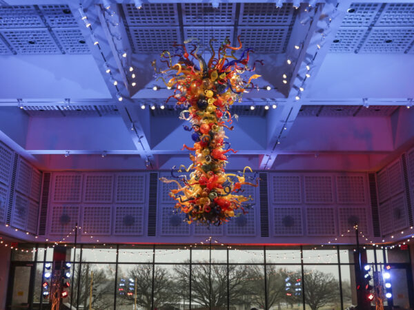 Photo of the colorful, large-scale Chihuly Chandelier in the S. Jim and Darla Farha Great Hall with small, white lights hanging from the ceiling around it and large windows in the background