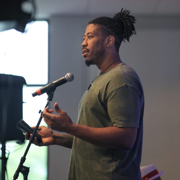 Photograph of African American man standing in front of a microphone, arms held in front of his torso with palms up. His dark hair is tied in a bun at the top of his head and he wears an olive green t-shirt