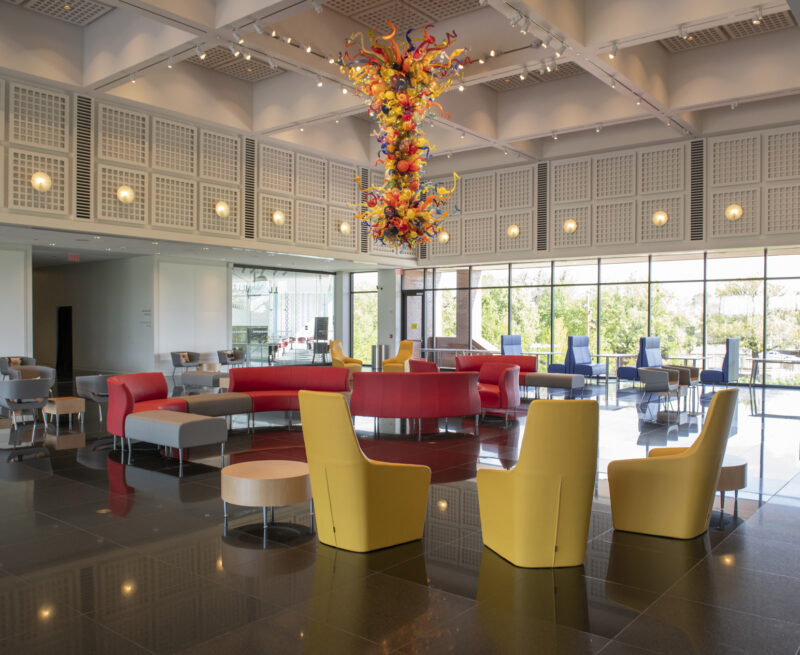 Photo of the S. Jim and Darla Farha with a dark granite floor, yellow chairs in the foreground, red couches in the middle and blue booths against the windows at the back of the hall with colorful glass Chihuly chandelier hanging from the ceiling