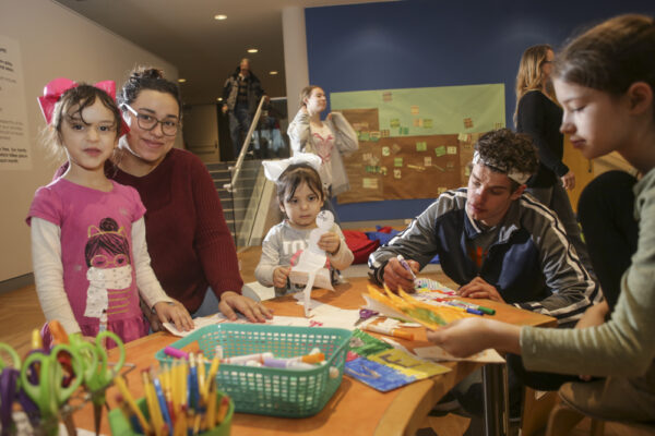 Photograph of people around a table with art supplies -- scissors, pencils, teal plastic basket with glue sticks and colorful drawings they have made. From left, dark-haired girl with a large pink bow on the back of her head and wearing a pink shirt with a face wrapped in a white scarf on the front; woman with dark hair and dark glasses, wearing a long-sleeved maroon shirt, in the center a younger girl holding a cutout snowman. She has dark hair with a large white bow and is wearing a gray sweater. Next to her is an adult male, dark-haired, wearing a black jacket with white stripe from shoulder to wrist, with a marker in his hand, coloring a piece of paper in front on him on the table. At far right is another dark-haired girl wearing a light, mossy green long-sleeved shirt. She is holding a yellow and green picture in her hand.