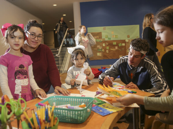 Photograph of people around a table with art supplies -- scissors, pencils, teal plastic basket with glue sticks and colorful drawings they have made. From left, dark-haired girl with a large pink bow on the back of her head and wearing a pink shirt with a face wrapped in a white scarf on the front; woman with dark hair and dark glasses, wearing a long-sleeved maroon shirt, in the center a younger girl holding a cutout snowman. She has dark hair with a large white bow and is wearing a gray sweater. Next to her is an adult male, dark-haired, wearing a black jacket with white stripe from shoulder to wrist, with a marker in his hand, coloring a piece of paper in front on him on the table. At far right is another dark-haired girl wearing a light, mossy green long-sleeved shirt. She is holding a yellow and green picture in her hand.