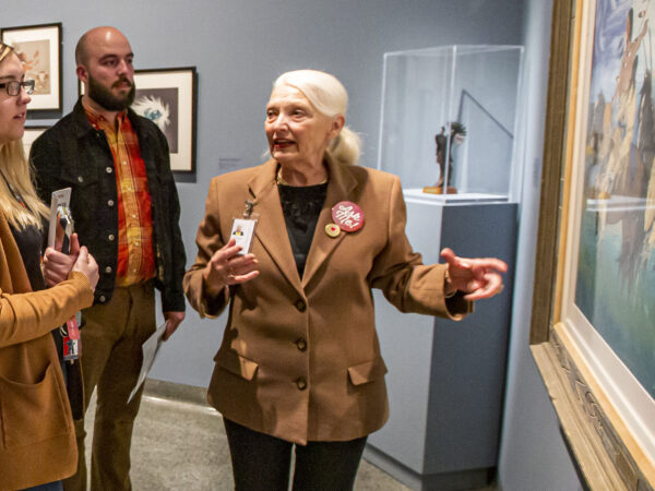 Photograph of people int the gallery. Woman at right has long blonde hair, dark-rimmed glasses and is wearing a tan coat. Man int he middle is bald with a dark beard, wearing an orange and yellow shirt under a dark jacket. Woman at right, standing in front of a painting, has white hair pulled back into a ponytail and is wearing a tan jacket over a black shirt. Her arm is outstretched with palm up toward the painting