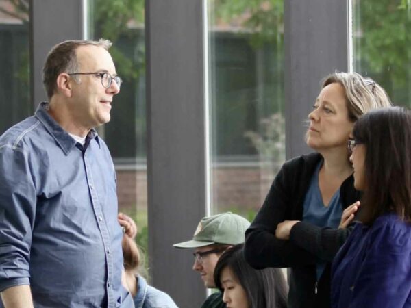 Photograph of a man and two women standing in front of floor-to-ceiling windows. The man has light brown hair and is wearing glasses, dressed in a blue, long-sleeved shirt with the cuffs rolled to his elbows. The woman in the center has light brown hair andis dessed in a dark sweater. The woman on the far right has shoulder length dark hair and is wearing a dark purple top.