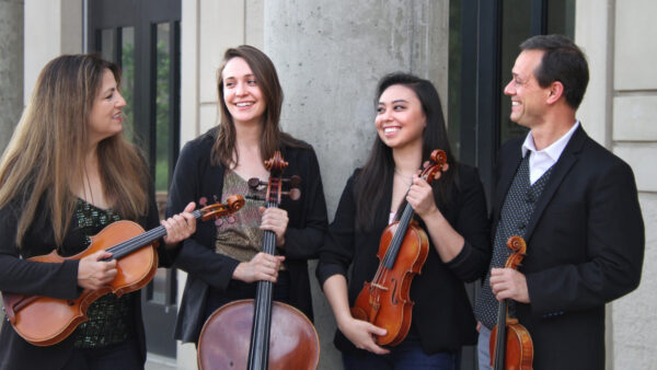 photograph of four people in an outdoor setting in front of a granite column. From left, woman with long dark hair, dressed in black and holding a viola, next is another woman with shoulder length dark hair, dressed in a black jacket over a lighter print and the fret and top of a cello in front of her. the third person is a woman with long, dark hair, dressed in black and holding a violin. The fourth person is a dark-haired man dressed ina black jacket and white shirt and holding a violin.