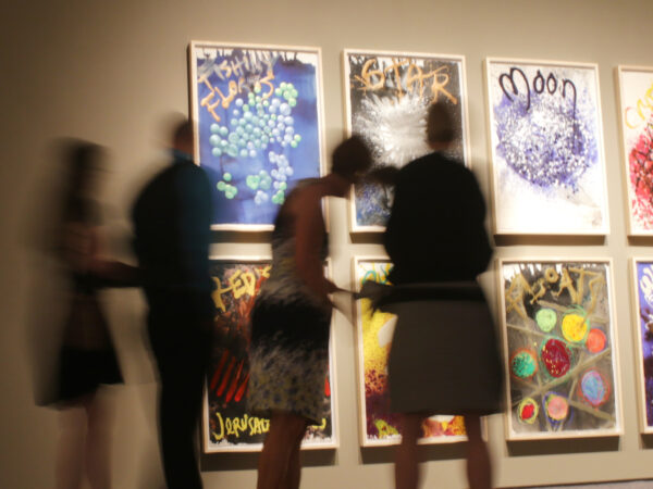 Photo of visitors blurry in the foreground and eight colorful prints on the wall in the background stacked in two rows and four columns