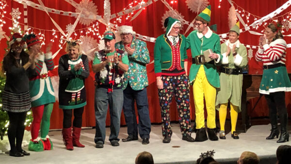 Nine people, four men and five women, stand on a stage and clap while wearing costumes inspired by the movie Elf