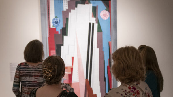 Photo of four women with the back of their heads toward the camera standing in front of a vertical abstract painting by Georgia O'Keeffe that has vertical lines of blue, white, pink, black, light brown, grey and coral colors
