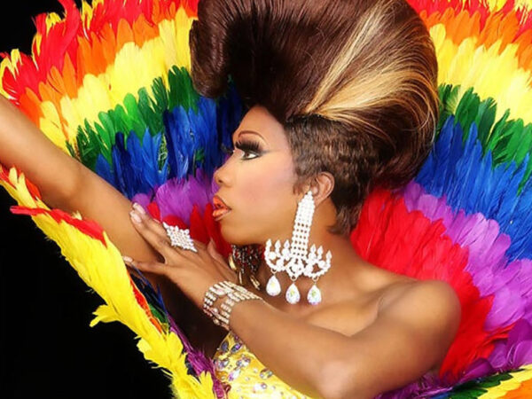 Profile of person with elaborate updo hairstyle against a colorful rainbow background. Arms are outstretched up to the left and she is wearing a long, glittery earring in an unside down T shape.