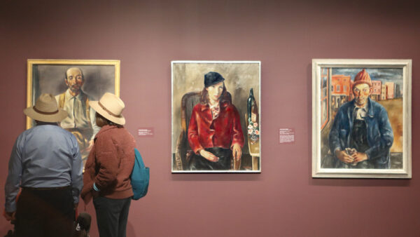 Two people standing in a gallery with three portraits on a lilght brown wall. The people, both seen from behind are wearing hats. The artwork they are standing in front of is obscured. To their left, the center artwork is a portrait of a seated women, wearing a bright red blouse and a black hat. The artwork on the far right is a seated man, wearing a denin-blue jacket and with a colorful landscape behind him.