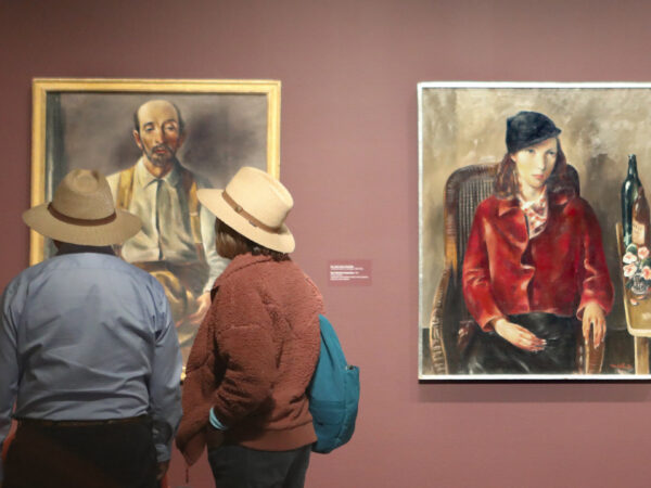 Two people standing in a gallery with three portraits on a lilght brown wall. The people, both seen from behind are wearing hats. The artwork they are standing in front of is obscured. To their left, the center artwork is a portrait of a seated women, wearing a bright red blouse and a black hat.