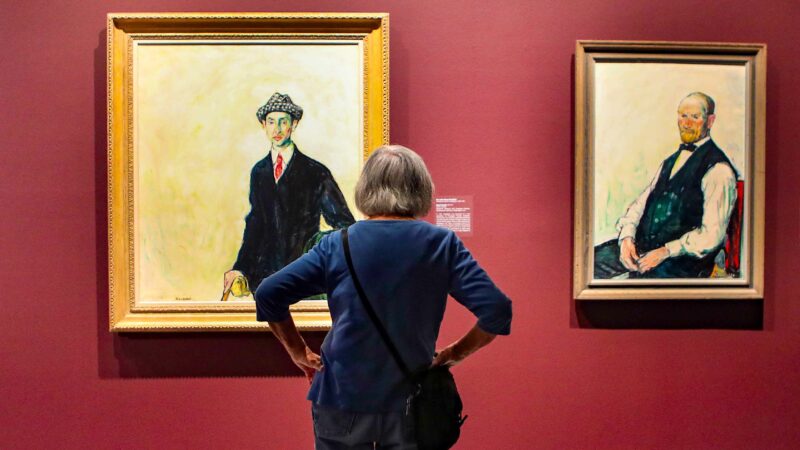 Woman pictured from the back standing with hands on hips in front of two portraits on men. Gallery wall is a red-orange color, portrait on the left is a man in a black suit, with a white shirt and red tie and wearing a dark hat on a pale yellow background.. Portrait on the right is a man sitting, wearing a black vest, and light colored shirt, on a pale yellow background white shi