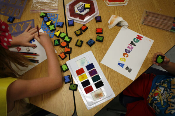 Color blocks of alphabetical letters on a table with an ink pad of six colors and two kids from the back, one on each side, showing their hands using the stamps and blocks to print on paper
