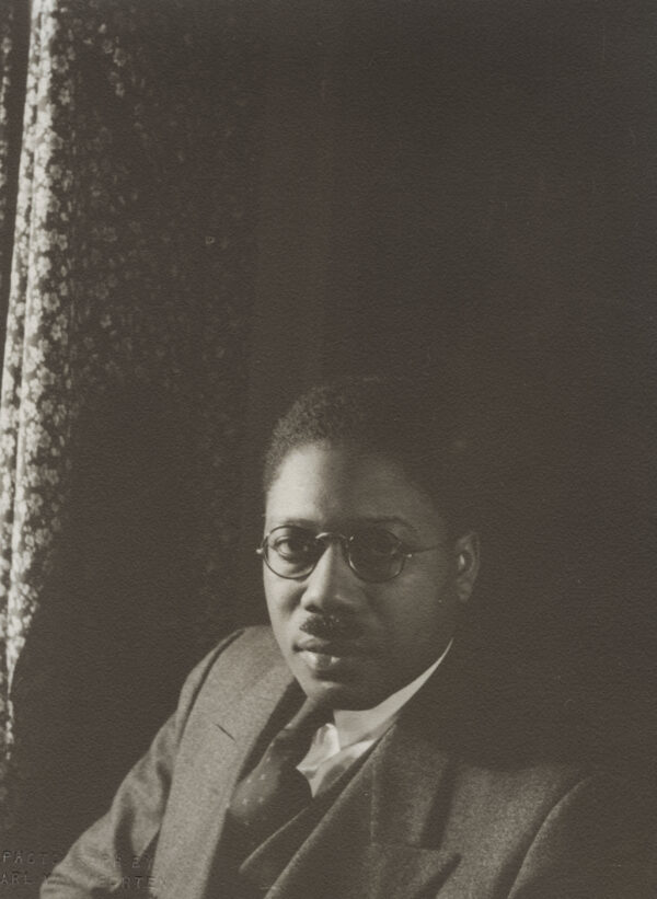Black and white photo of African American man with close-cropped hair, wearing glasses, and a suit, against a dark background, with light comeing from the left of the photo. There is a sliver of a print curtain at the left of the image.