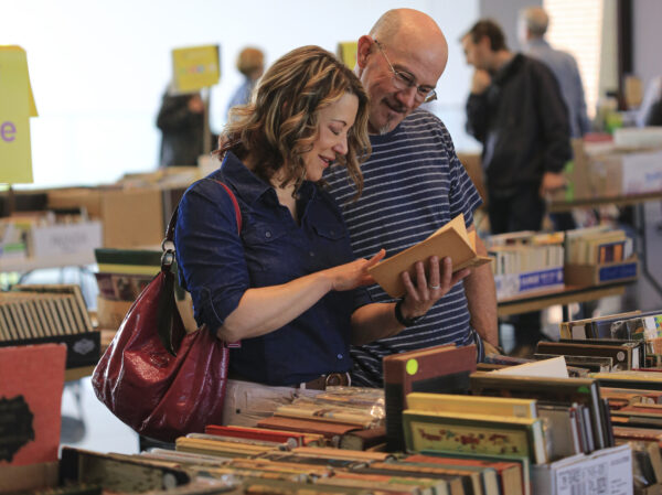 Couple standing in front of a tablecovered with books. Woman on the left holds a book in her hand and is pointing to a page inside the book. She is dressed in a blue button blouse and has a red leather bag over her right shoulder. The man looks over her left shoulder at the book. He is balding, wears glasses and and a blue and white striped shirt.