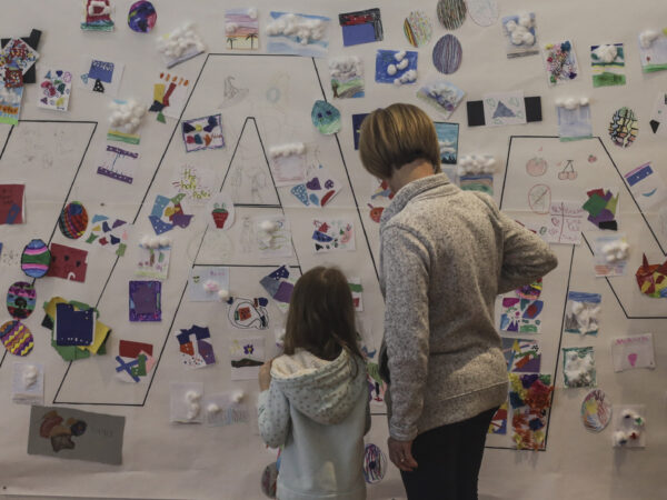 Young girl and woman with backs the the camera, looking at a white wall covered with notes and small pictures